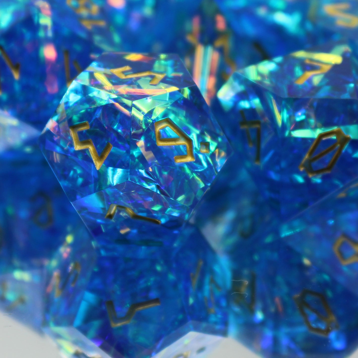 Close up of StarGlow blue dice arranged in a pile on a reflective surface. the D10 die is in focus, showcasing the gold numbers and iridescent material within the die.