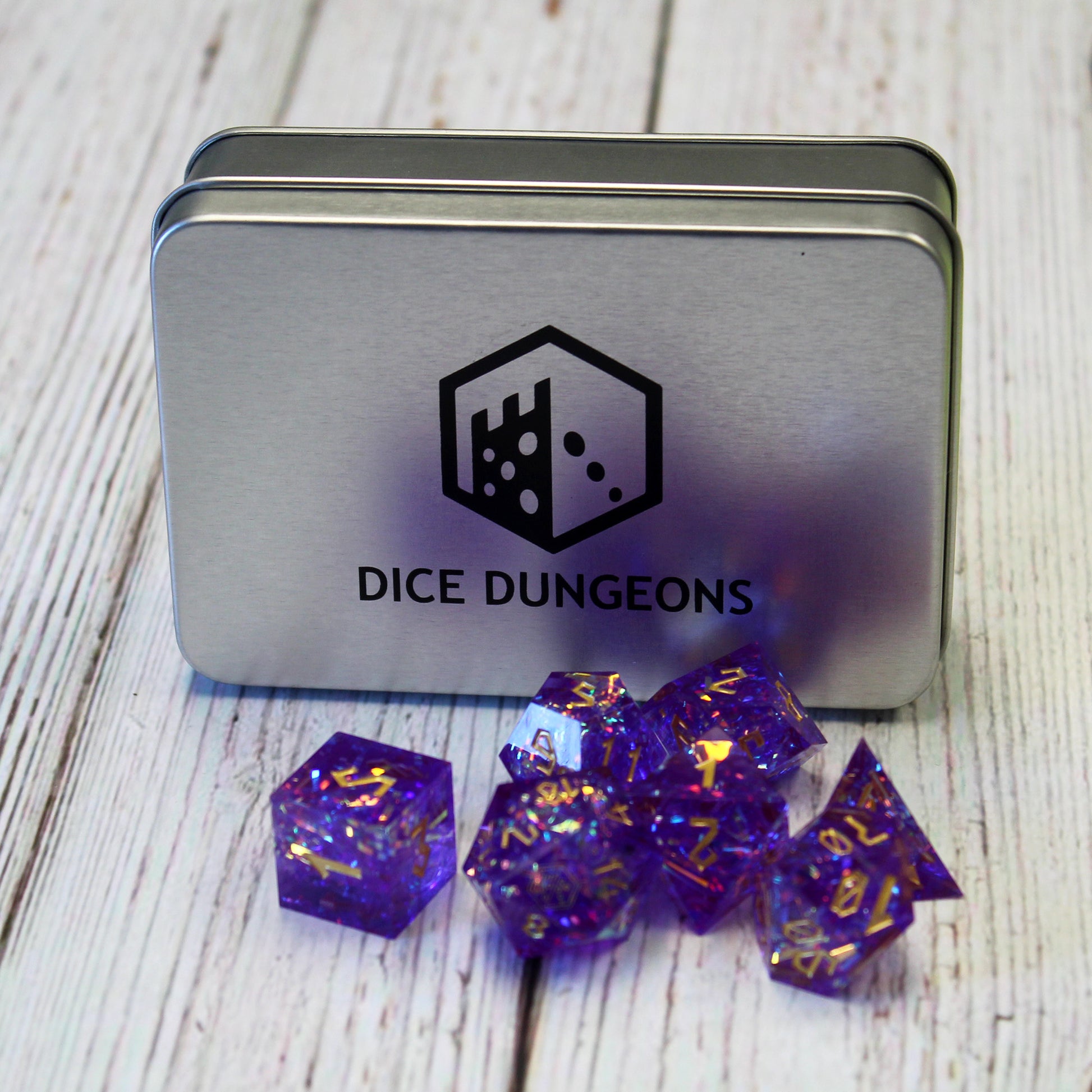 StarGlow Purple dice set arranged in a pile on a white wood surface. Behind the pile is a Dice Dungeons display box, a silver metal box with the Dice Dungeons logo printed in black on the front.