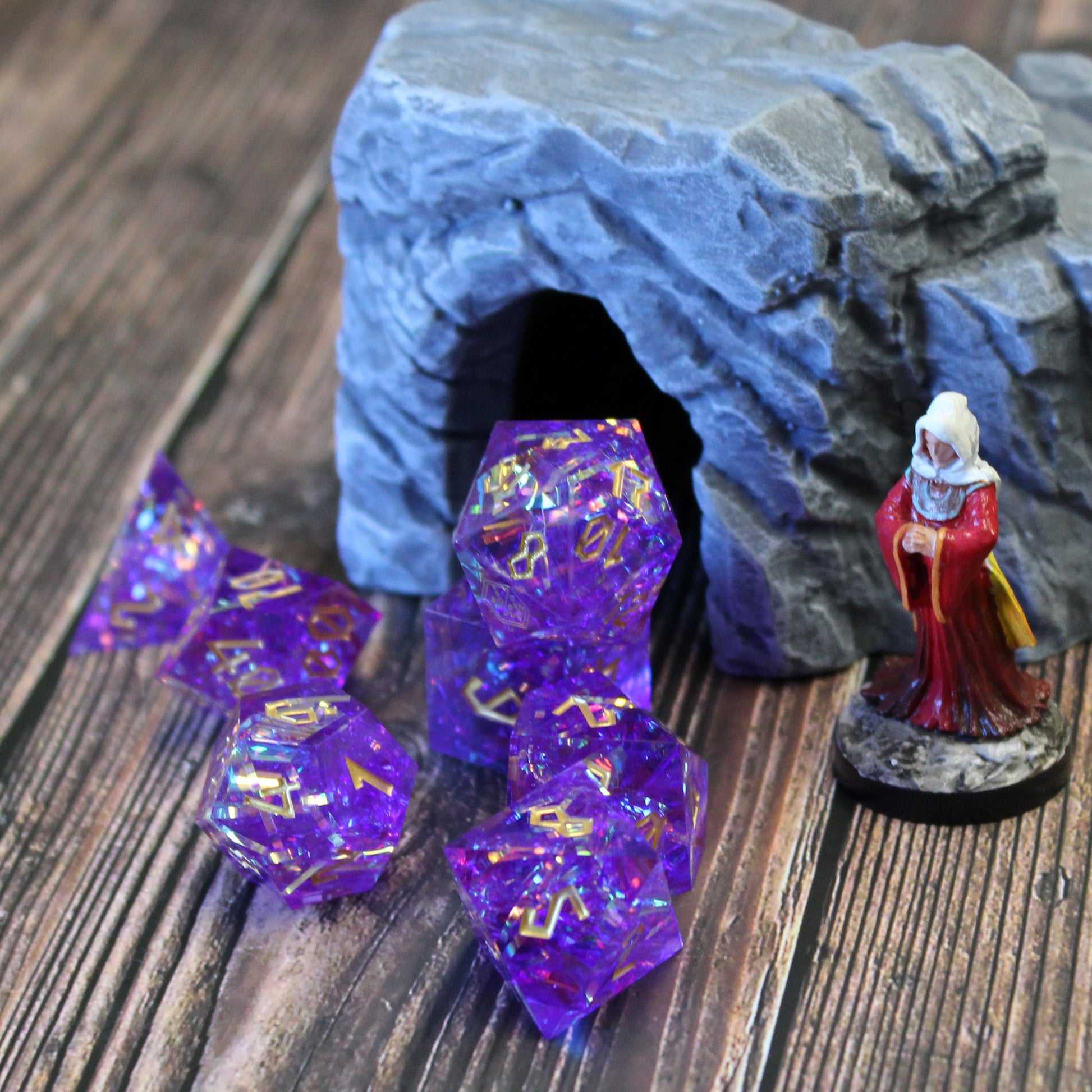StarGlow Purple dice set arranged in a pile on a wood surface. Next to the dice on the right is a mini figure, and behind the dice is a gray cave opening set piece that the dice have been arranged in the mouth of.