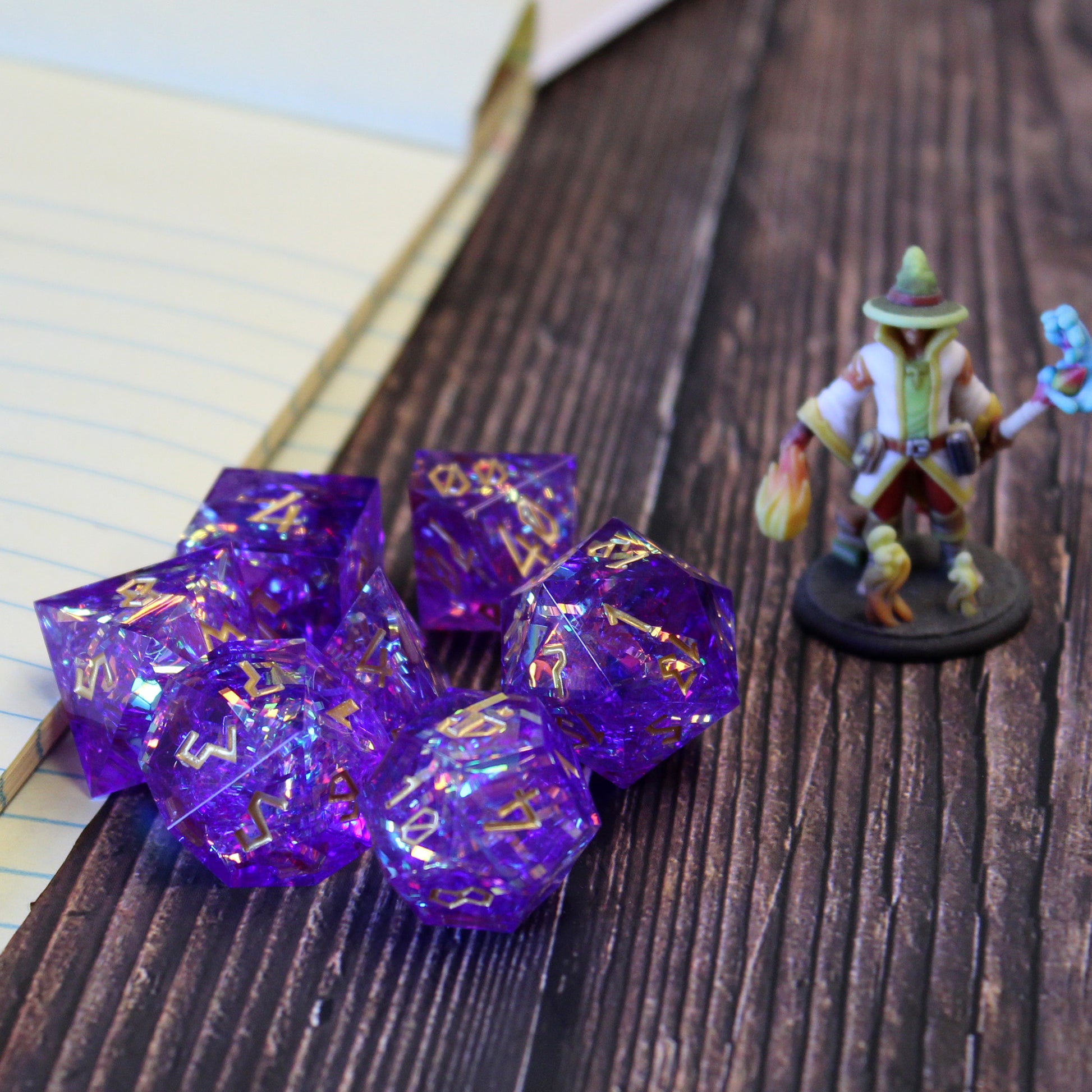 StarGlow Purple dice set arranged in a pile on a dark wood surface. To the right of the dice pile is a mini figure, and to the left is a stack of lined paper.