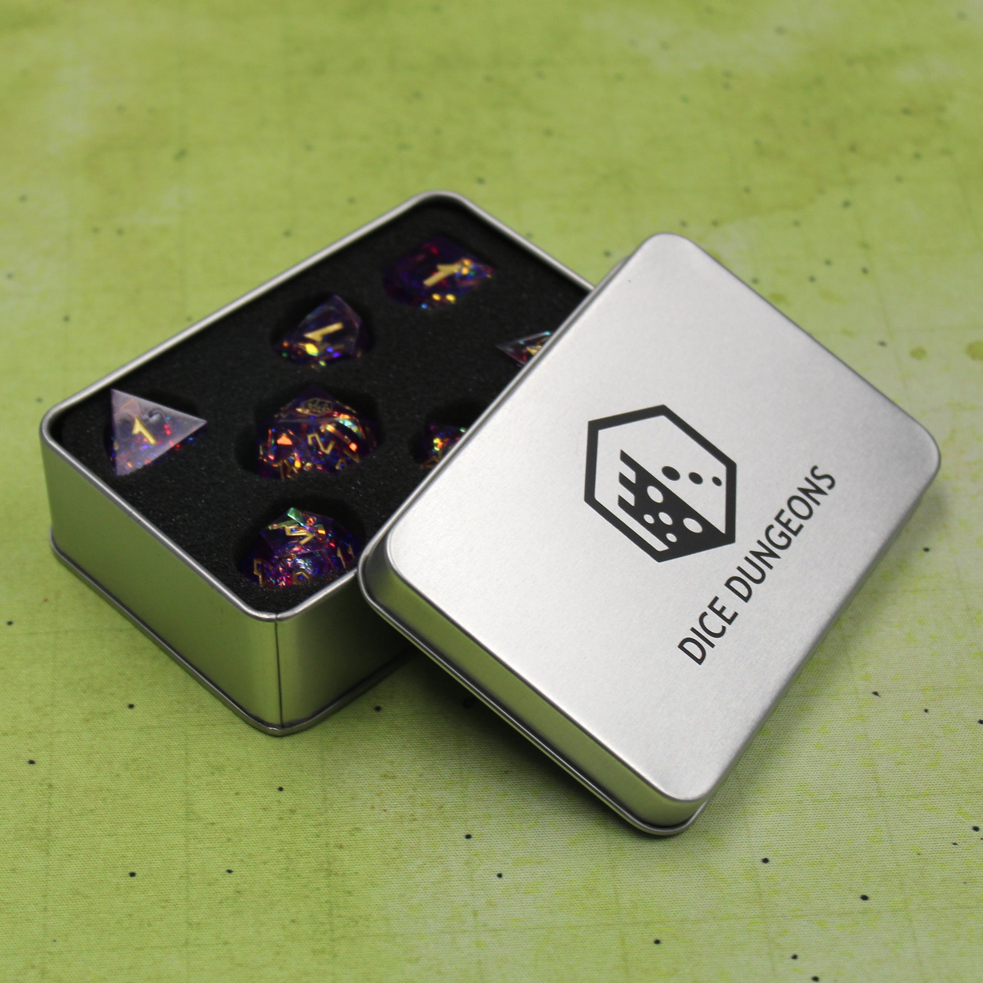 StarGlow Purple dice set stored within a Dice Dungeons dice box on a green game map. The dice box is open so that the individual spots for each die can be seen. The lid of the box is propped up to the side, and the black Dice Dungeons logo can be seen printed on the center of the silver metal box.