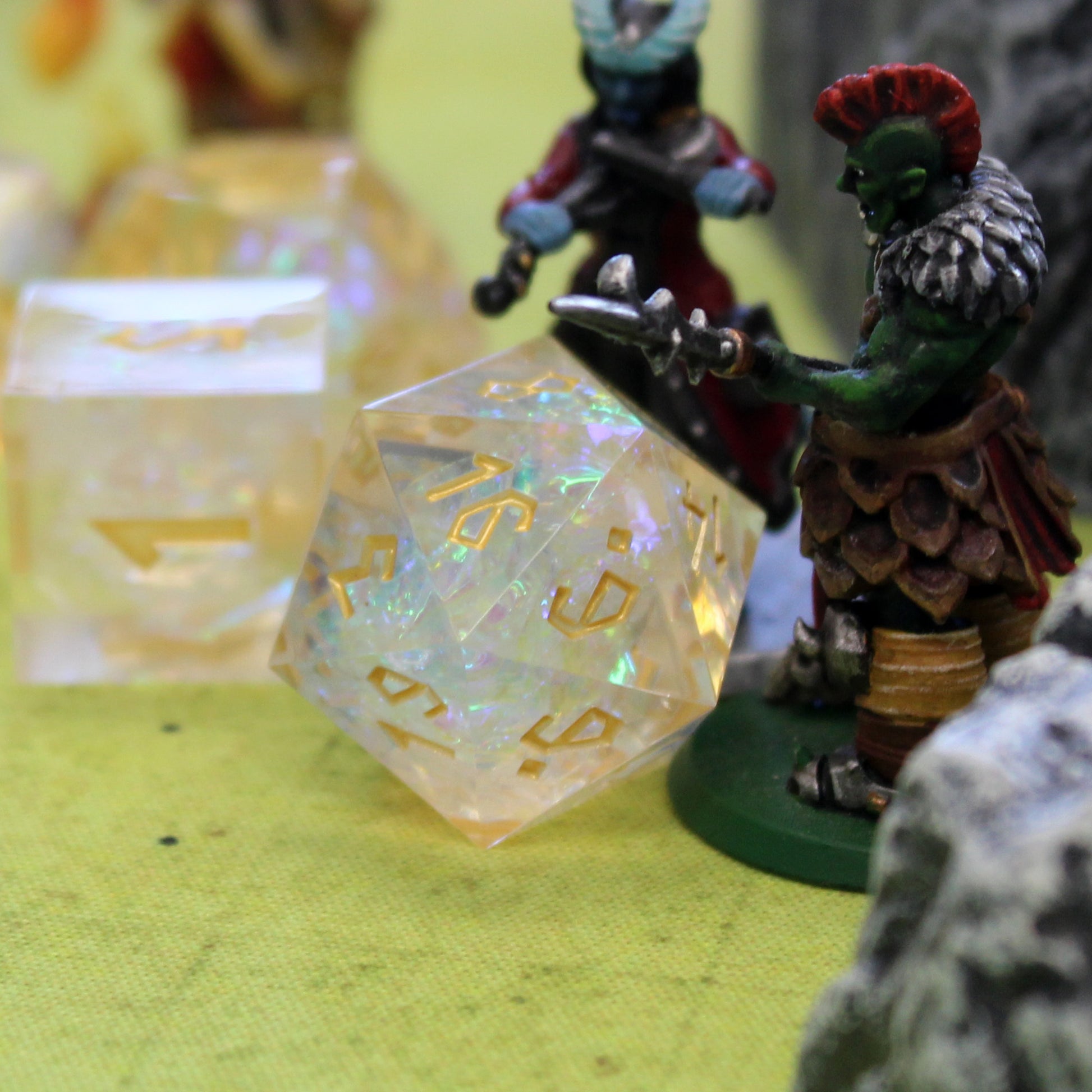 Upclose D20 with a few other dice scattered behind it and out of focus. The D20 has one mini figure to the right of out and one mini figure behind it. Everything is arranged on a green game mat.