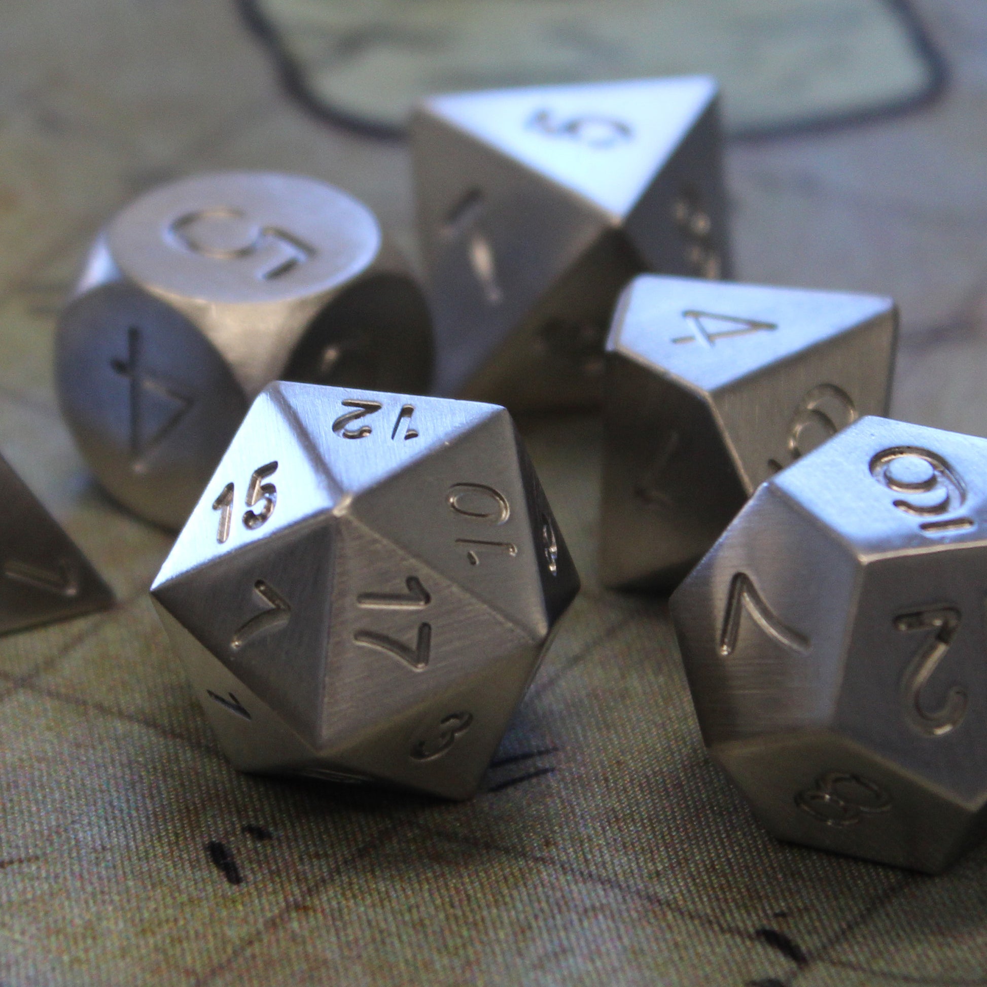 Close up photo of the Tungsten dice set resting on a 1 inch map grid. The d20 is front and center with the 12 face up and clear detailing of the brushed metal surface and engraved numbering of this unplated set.