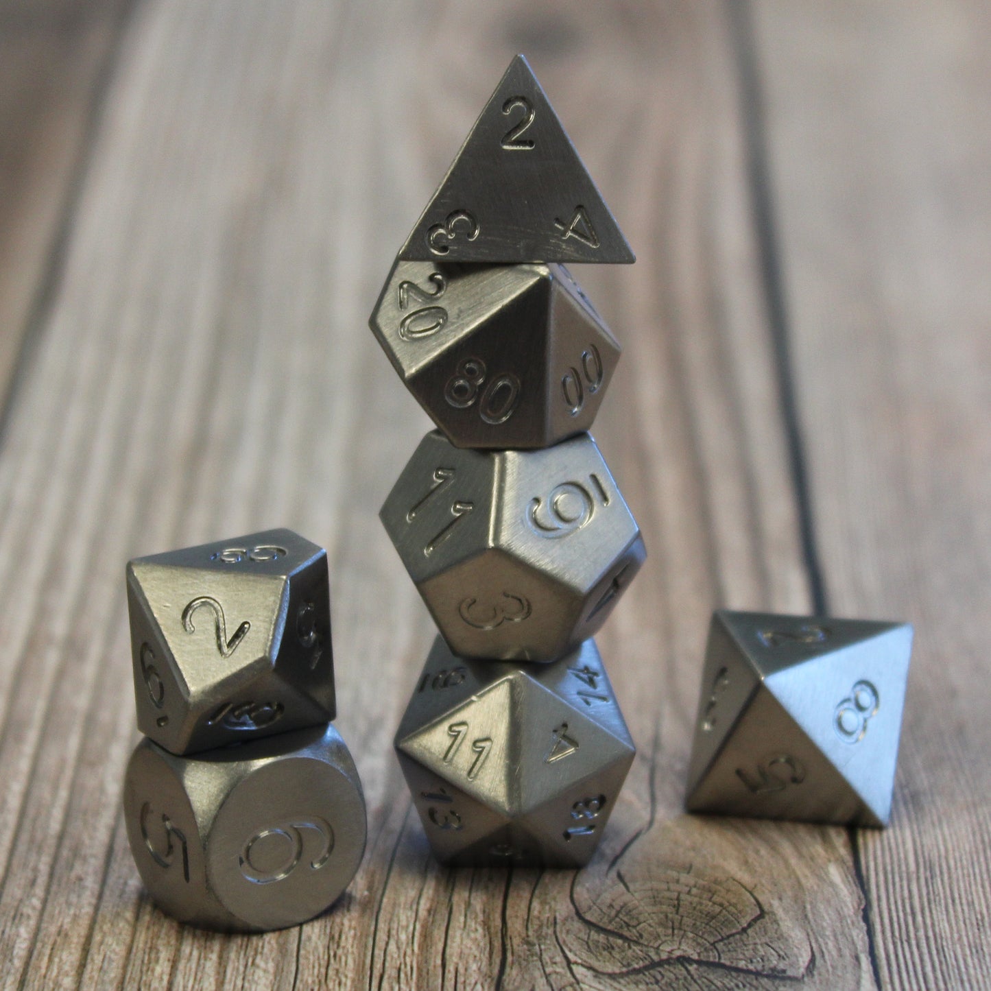 Metal Tungsten RPG dice set arranged in stacked columns on a wooden surface. This set has no electroplating or number inking and appears as a greyish-white lustrous metal.