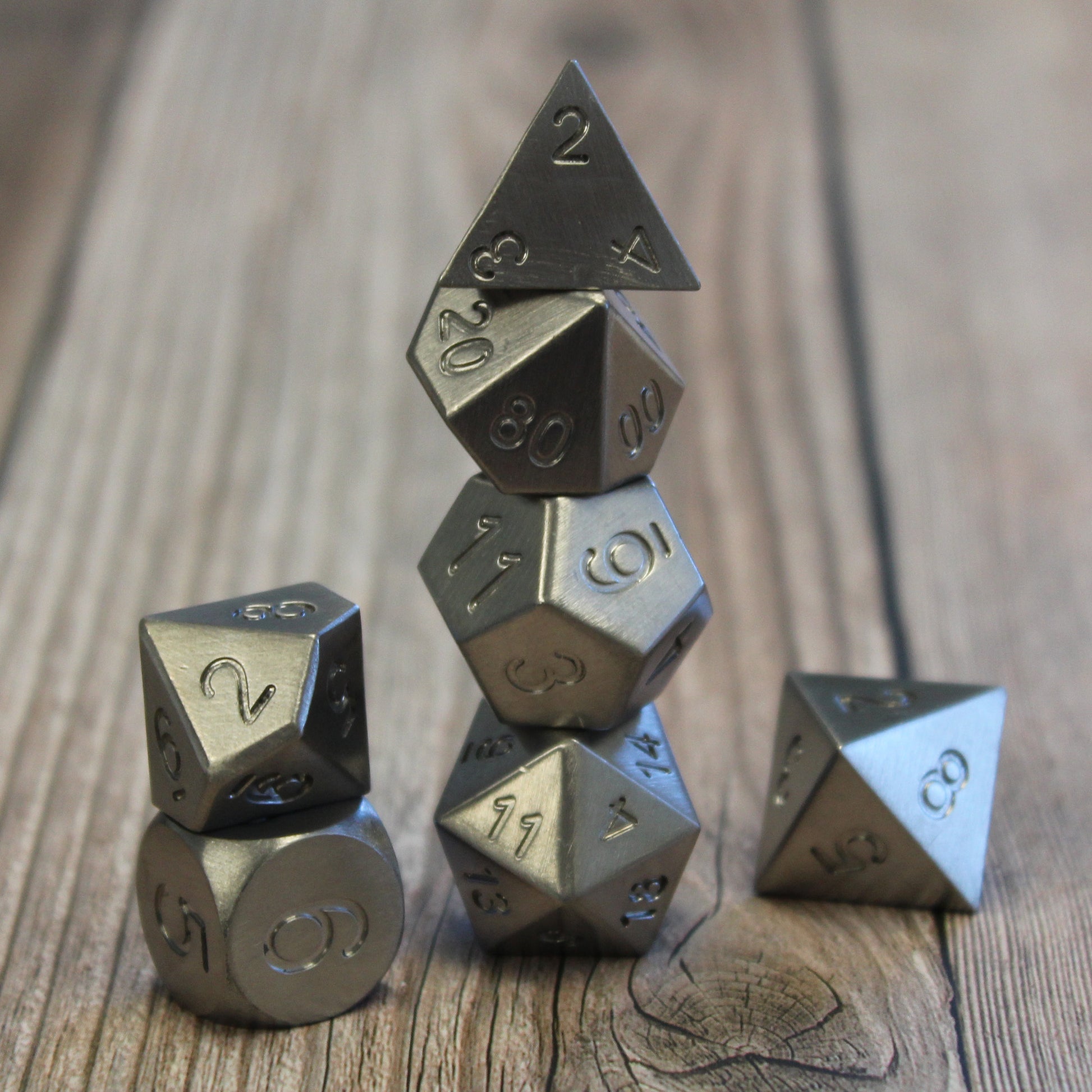 Metal Tungsten RPG dice set arranged in stacked columns on a wooden surface.  This set has no electroplating or number inking and appears as a greyish-white lustrous metal.