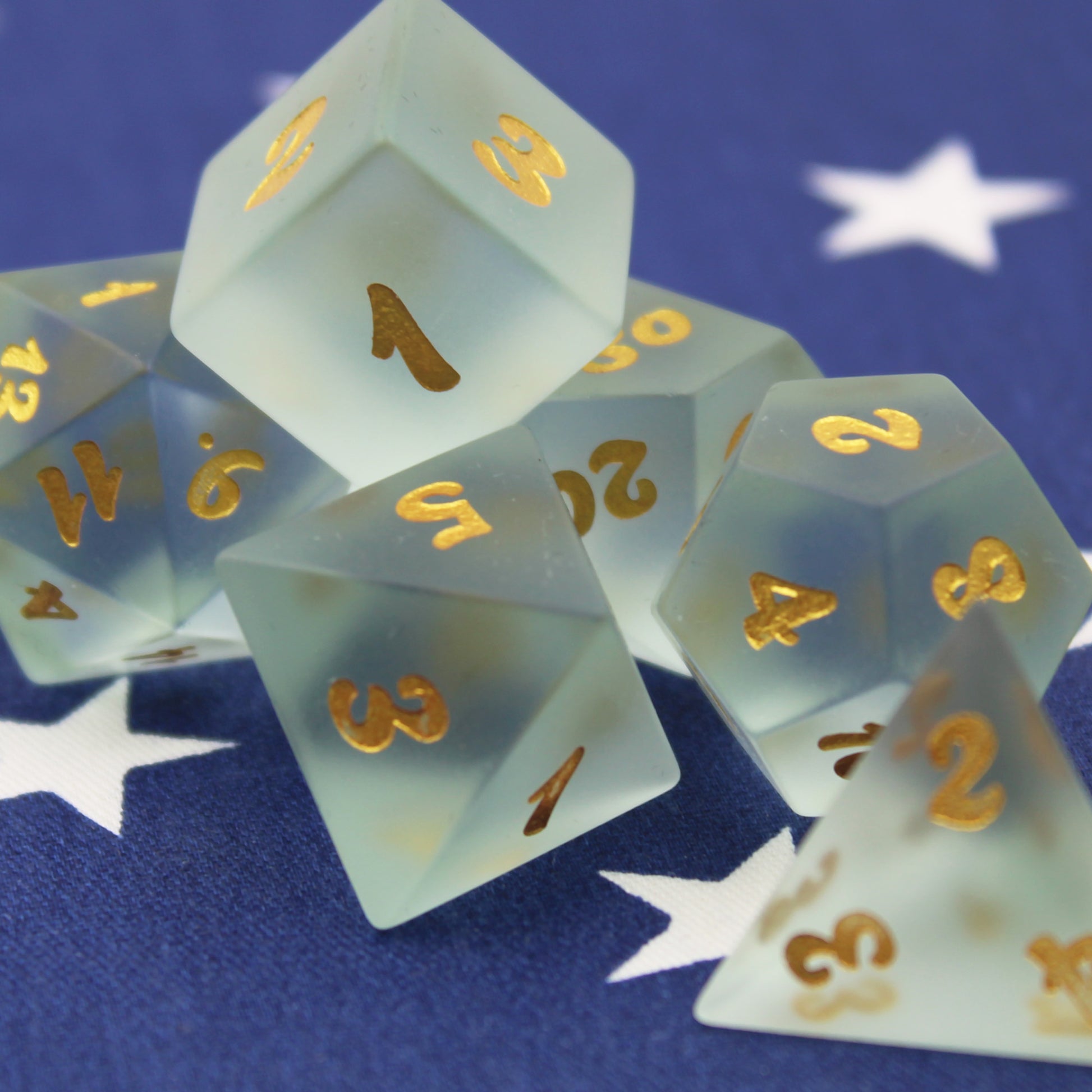 Close pp of powder blue frosted glass dice with gold numbering on a blue fabric background.