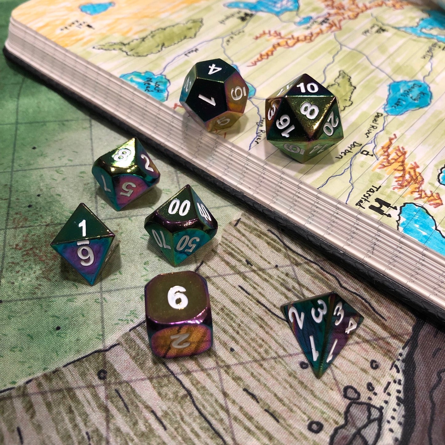 Alchemist's Oil dice arranged on top of a printed map surface and an open notebook with a detailed map drawn inside.