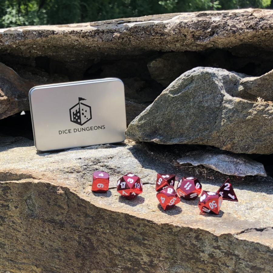 Radiant red dice set scattered on a stone outside with a Dice Dungeons silver display box propped up behind them as well as other large stones.