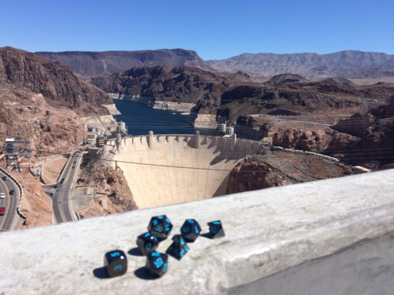 Metal Black Frost dice set arranged on a ledge overlooking the Hoover Dam with red mountains behind