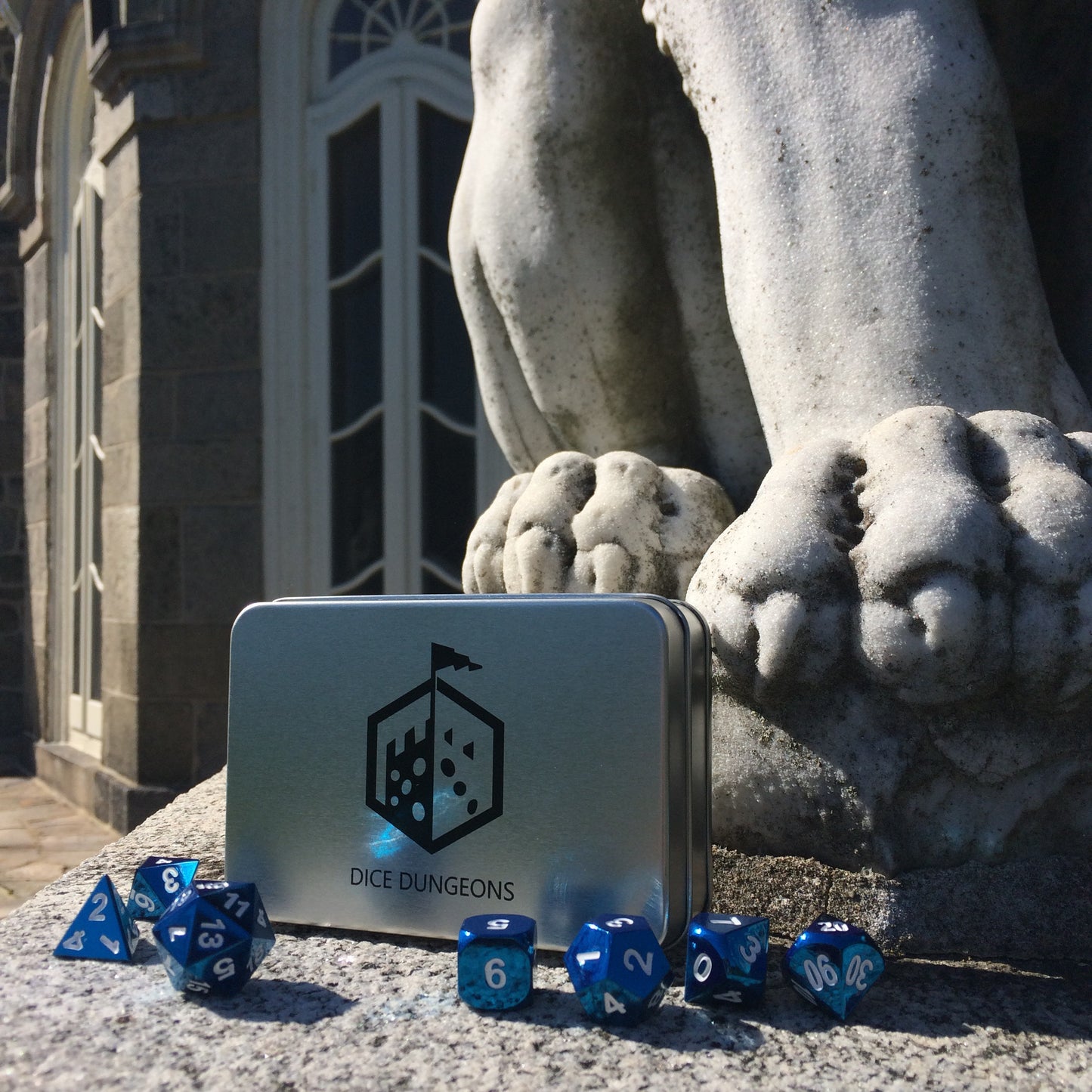 Radiant blue dice set scattered in front of a Dice Dungeons silver display box on a stony floor. Behind them are a mythical creature and castle set piece.