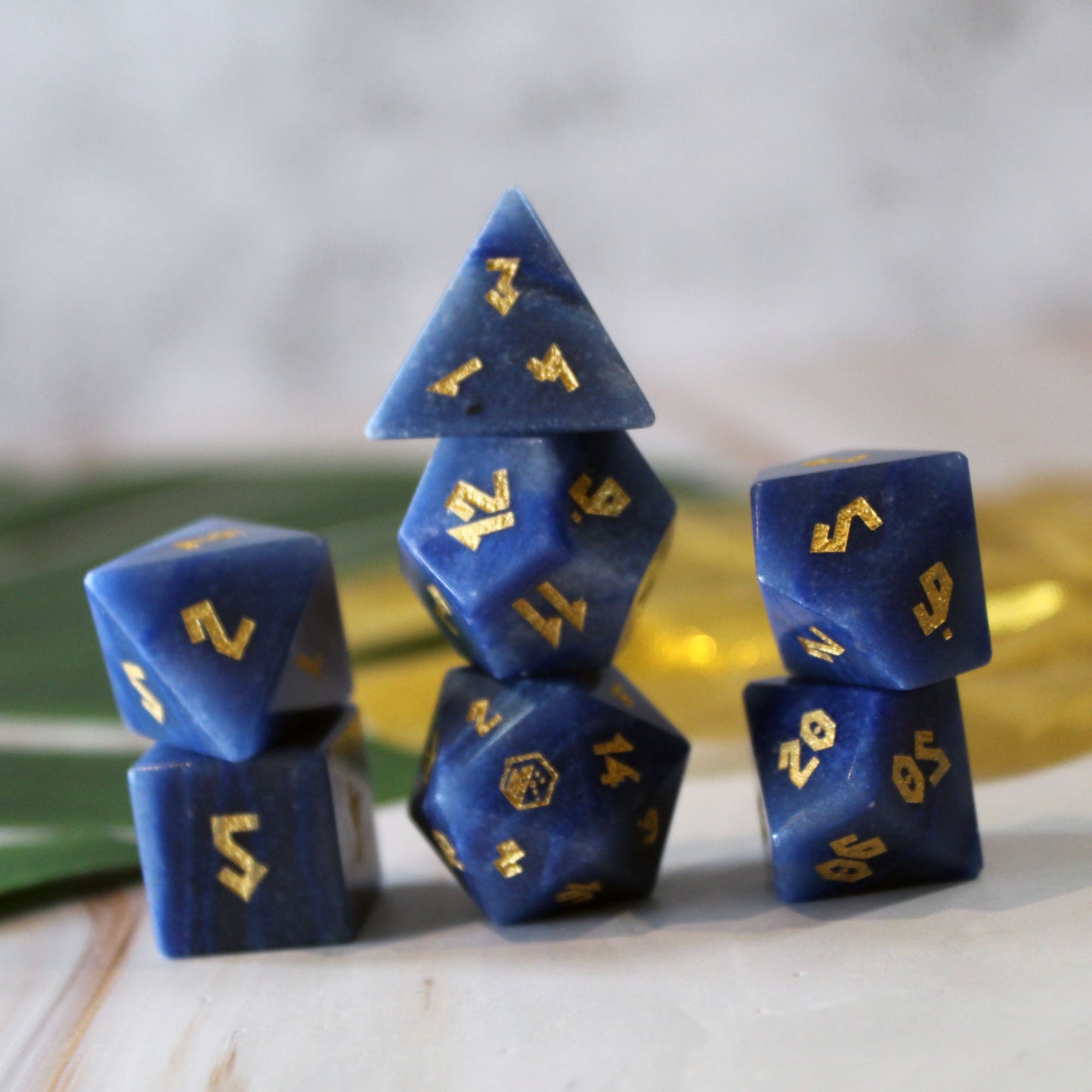 A set of blue gemstone gaming dice with gold numbers, arranged in a stack on a light marble-like surface with a soft blurred background.