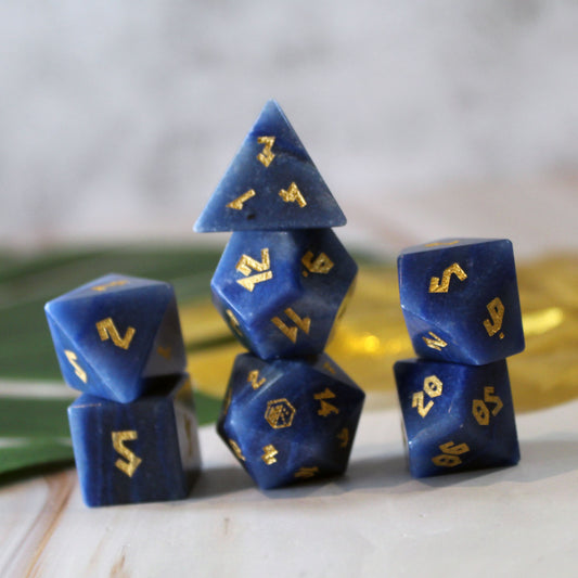 A set of blue gemstone gaming dice with gold numbers, arranged in a stack on a light marble-like surface with a soft blurred background.