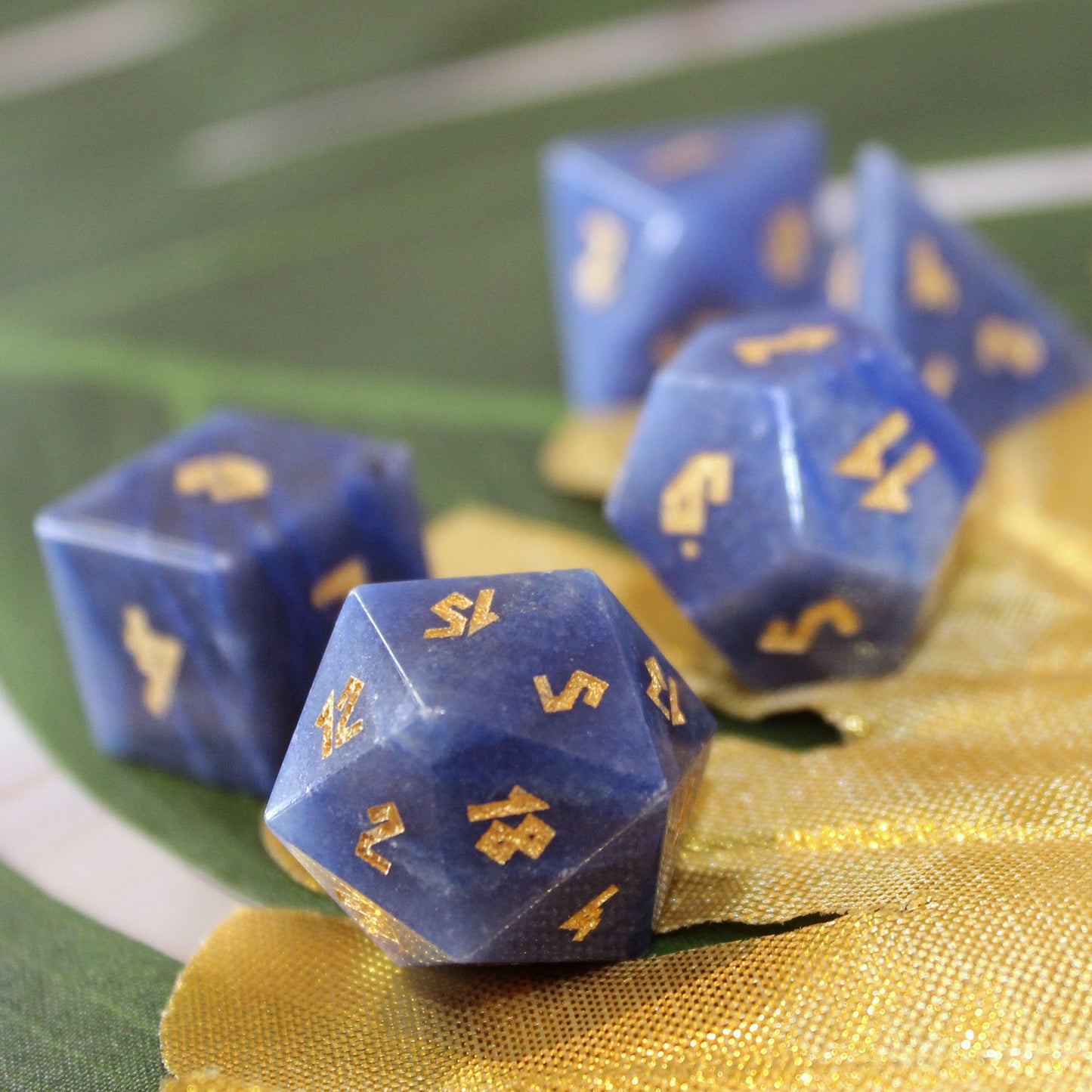 Blue Adventurine polyhedral tabletop gaming dice with gold numbers, arranged on a gold fabric surface with a blurred green and white background.
