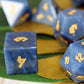 Blue gemstone polyhedral dice for dungeons and dragons with gold-colored numbering, shown in a close-up shot with a green and gold striped fabric background.