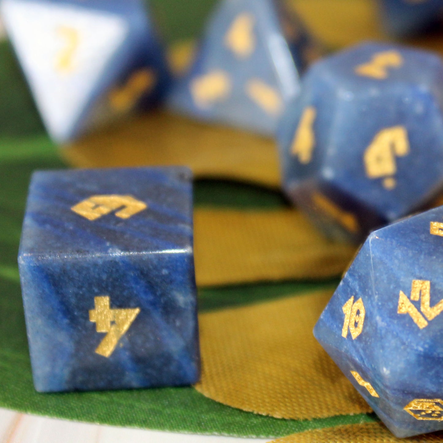Blue gemstone polyhedral dice for dungeons and dragons with gold-colored numbering, shown in a close-up shot with a green and gold striped fabric background.