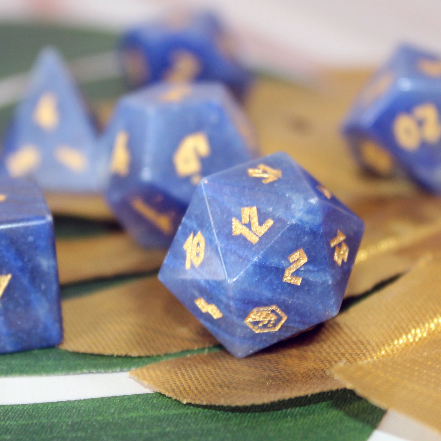 Blue polyhedral tabletop gaming dice made from the gemstone Blue Adventurine with gold-colored numbers, arranged on a green and gold textured fabric surface.