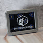 A square silver metal tin with a clear front window displaying blue polyhedral dice inside. The tin has a white 'DICE DUNGEONS' logo and text on a black background, resting on a light marble surface against a textured gray wall.