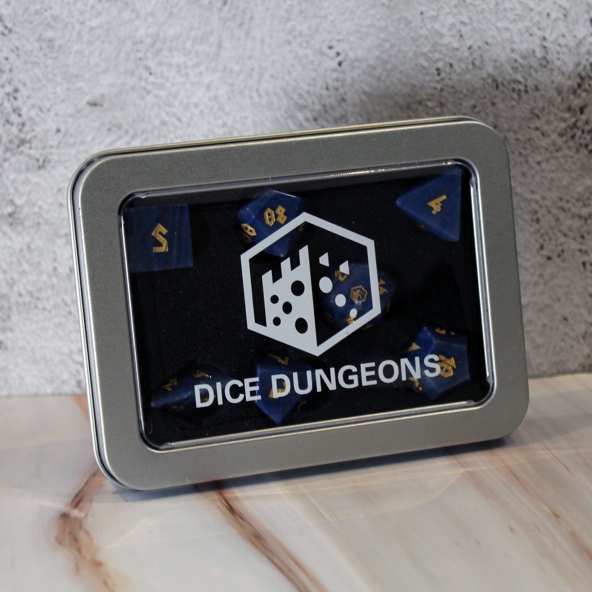 A square silver metal tin with a clear front window displaying blue polyhedral dice inside. The tin has a white 'DICE DUNGEONS' logo and text on a black background, resting on a light marble surface against a textured gray wall.