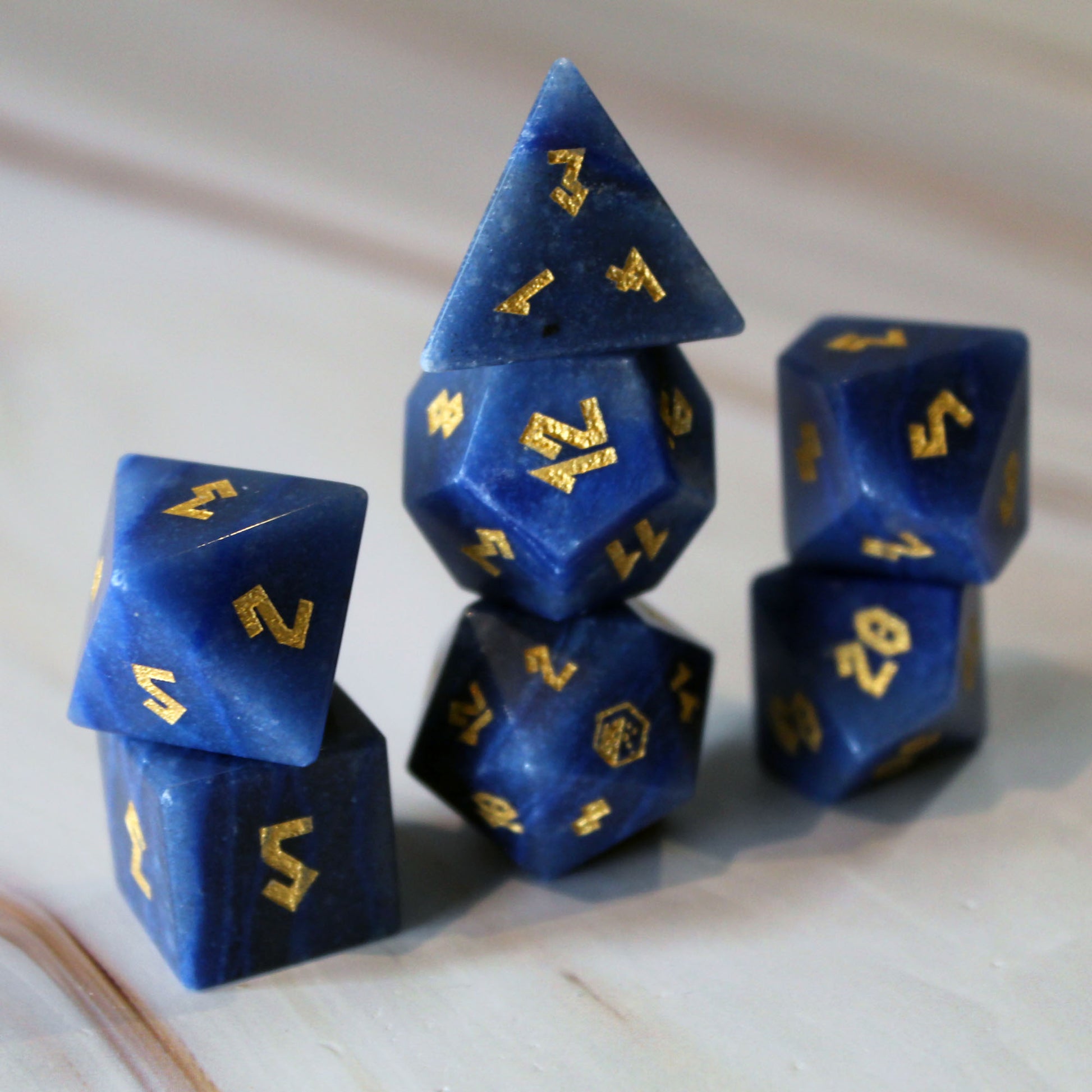 A set of blue gemstone polyhedral dice with gold-colored numbers, arranged on a light-colored wooden surface.