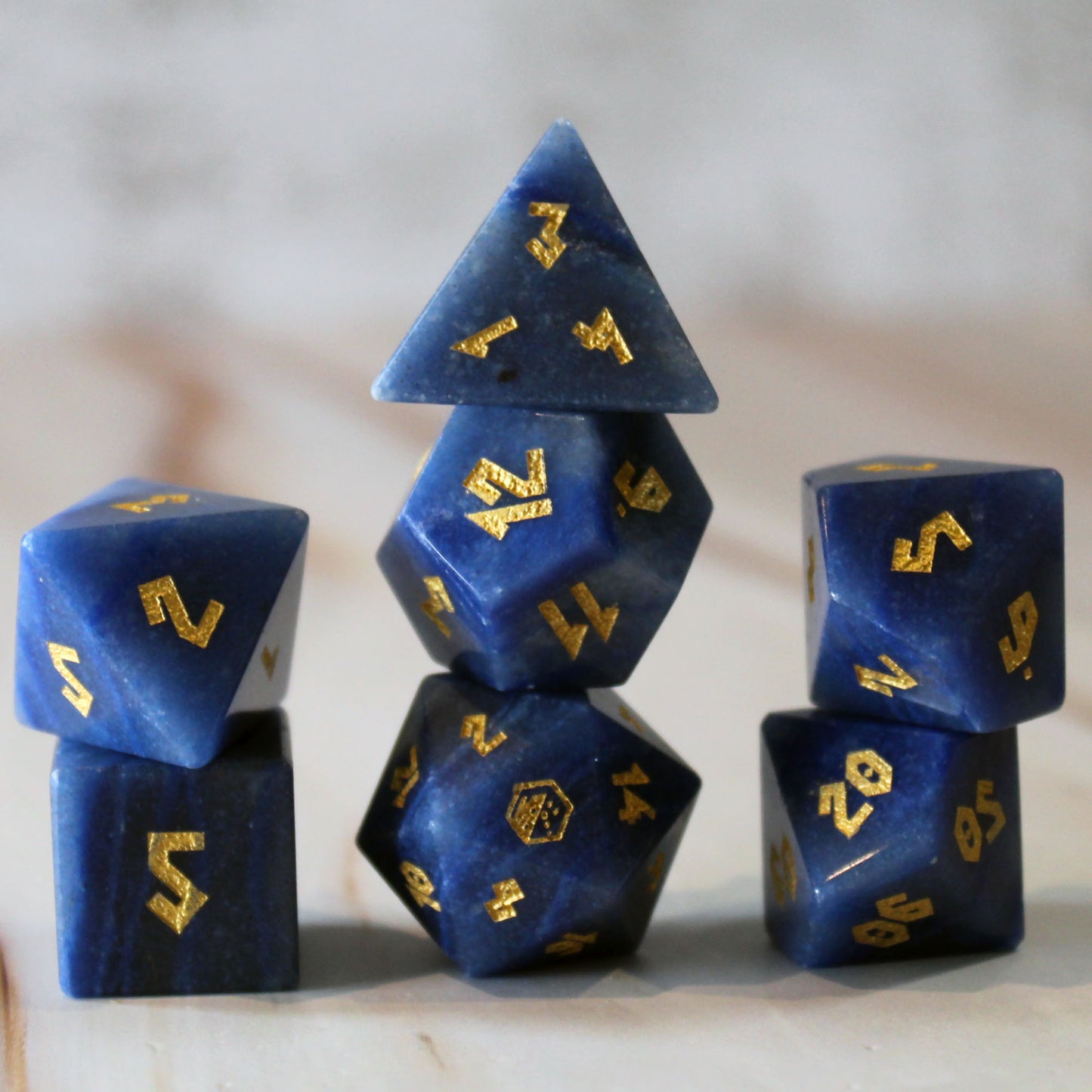 A set of blue polyhedral gaming dice with shiny gold numbers, arranged in a stack on a light marble-like surface. The set is made from the gemstone Blue Adventurine. 