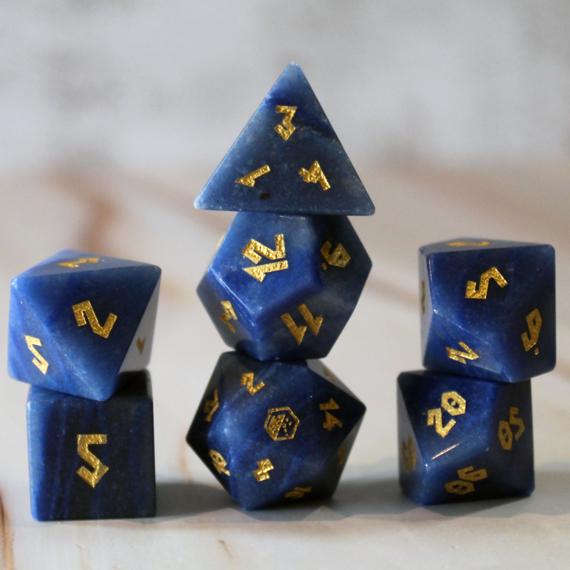 A set of blue polyhedral gaming dice with shiny gold numbers, arranged in a stack on a light marble-like surface. The set is made from the gemstone Blue Adventurine. 