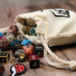 A full cotton dice bag laying down with a variety of polyhedral dice spilling from its opening.