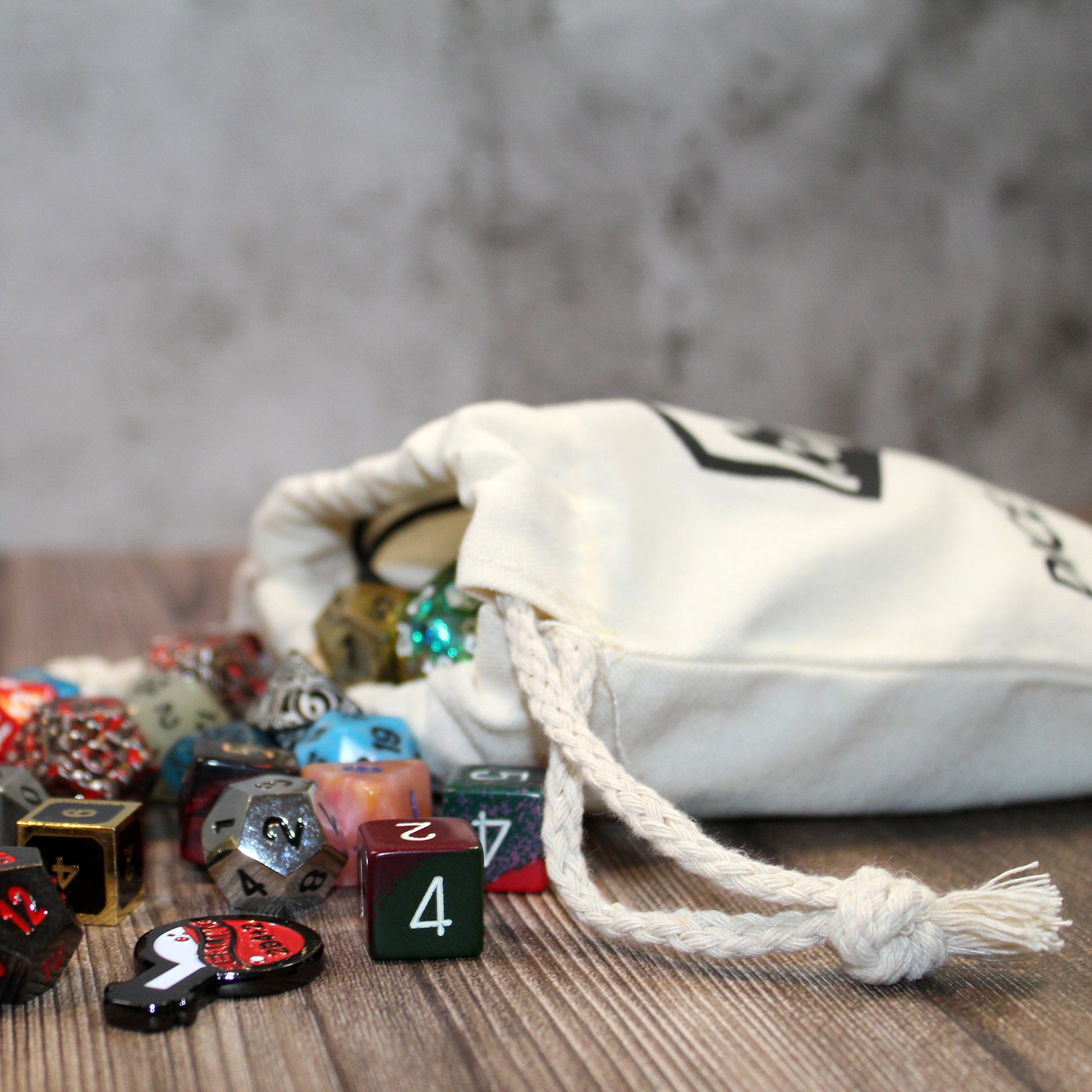 The side view of a full cotton dice bag laying down with a variety of polyhedral dice spilling from its opening.