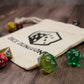 An empty cotton dice bag laying flat on a wooden surface with a few dice scattered around it.
