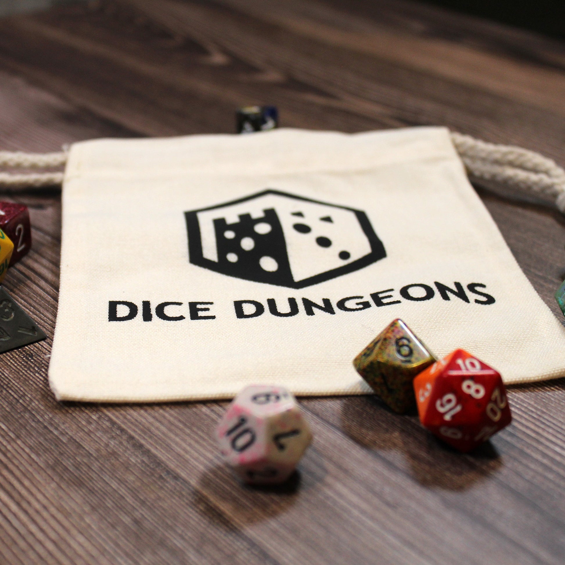 An empty cotton dice bag laying flat on a table with "Dice Dungeons" visible on its front and a few polyhedral dice scattered around it.