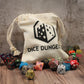 A full cotton dice bag sitting upright on a wooden surface with a collection of polyhedral dice scattered around it.