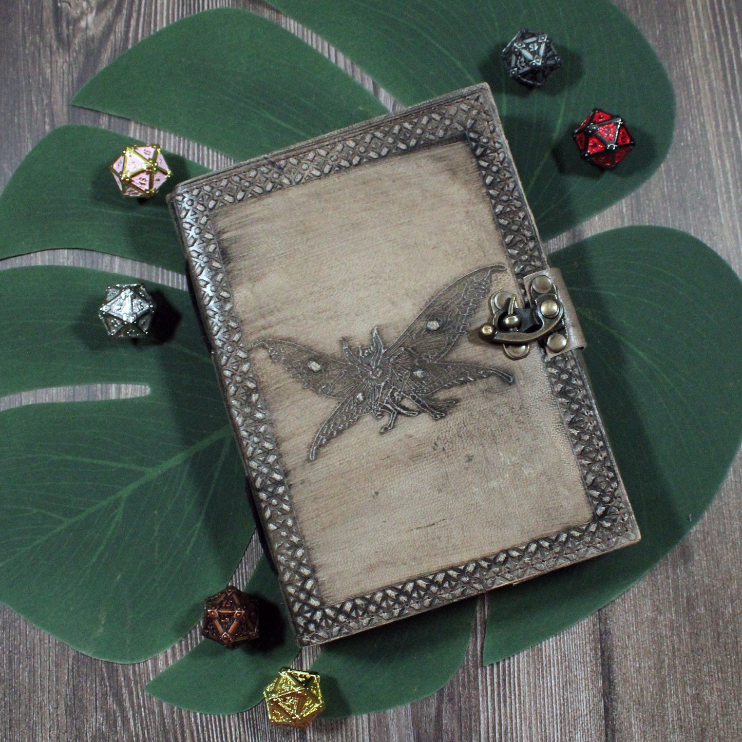 Decorative notebook with a moth-man design on a wooden surface with dice around it. Used for dungeons and dragons and tabletop game notes.