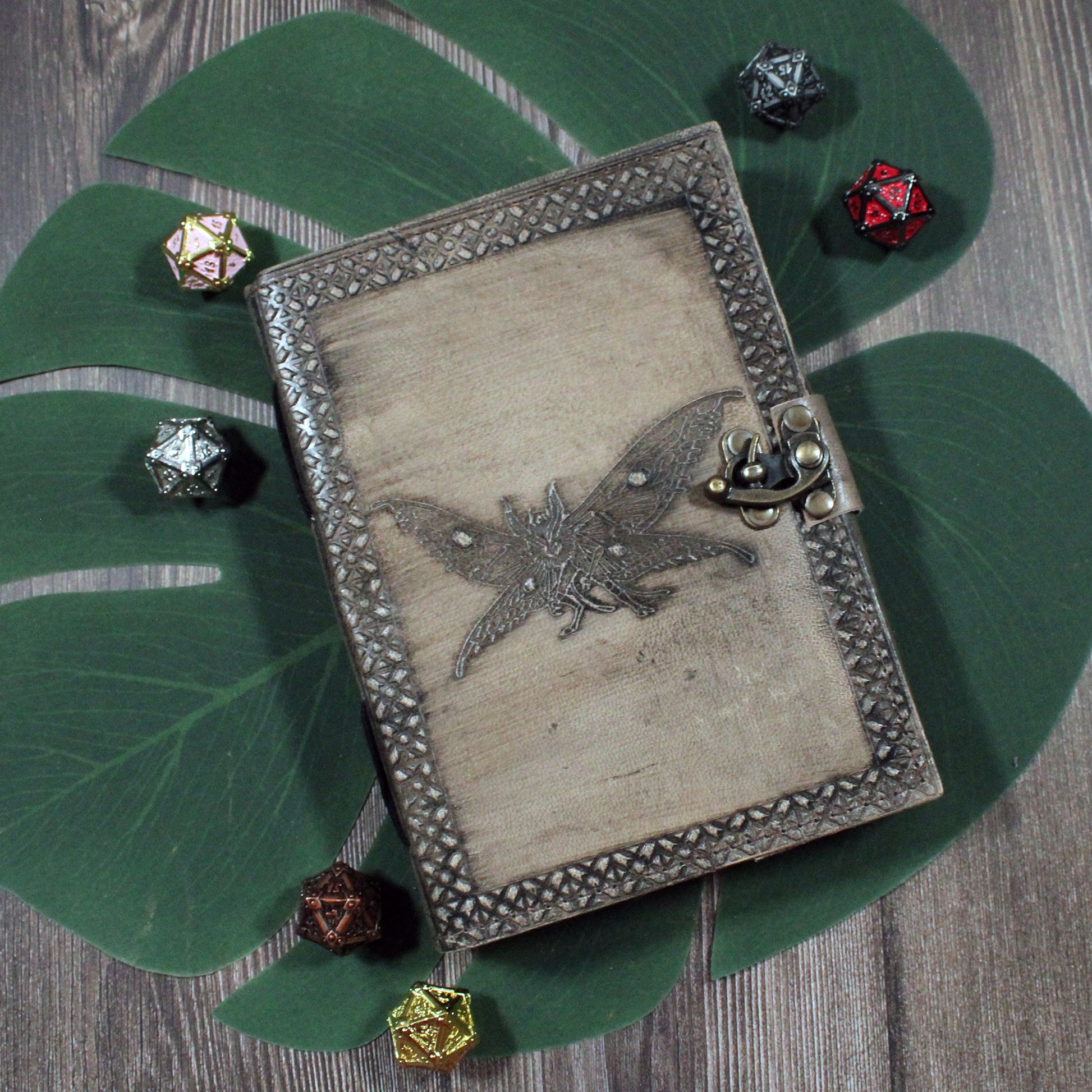 Decorative notebook with a moth-man design on a wooden surface with dice around it. Used for dungeons and dragons and tabletop game notes.