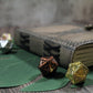 Two metallic dice, one gold and one bronze, on green leaves with a leather journal in the background.