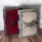 Two ornate leather-bound journals with embossed designs of the cryptid mothman on a wooden surface.