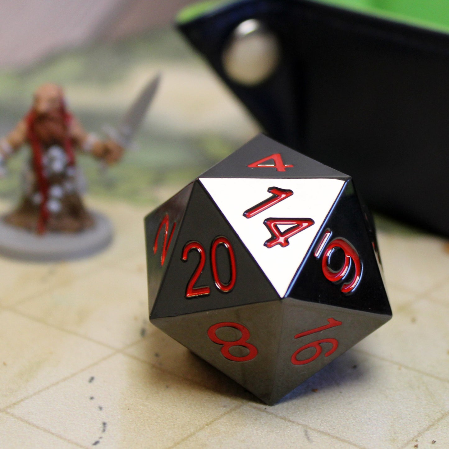 Jumbo 33mm Metal D20 Ember Dice on Game Table with Miniature for Scale, Black Nickel-Zinc Finish with Red Numbering, Perfect for RPGs.