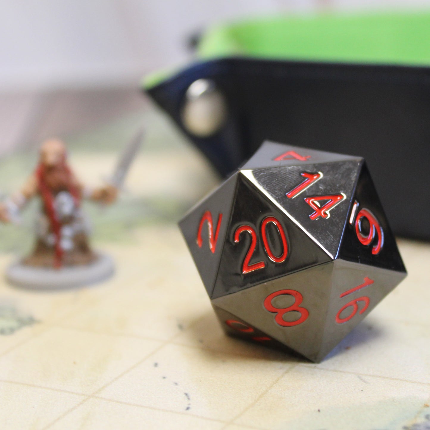 Jumbo Metal Ember D20 on Dungeons & Dragons Battlemap next to a figurine for size and in front of a green dice tray, Featuring Black Nickel-Zinc Finish and Red Numbering.