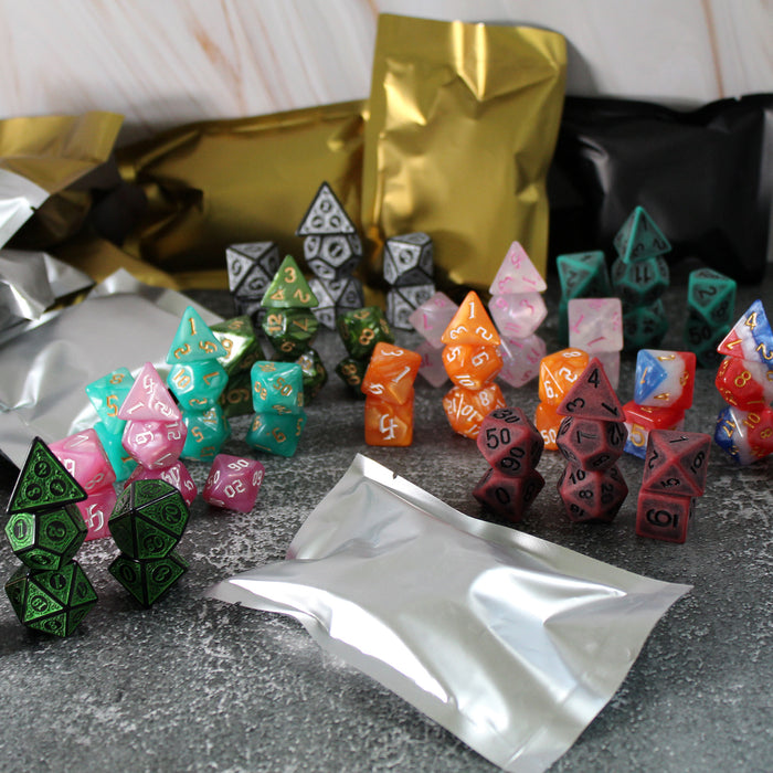 Stacks of different polymer dice sets of varying bright colors with one silver foil bag resting in front of them, on a textured gray surface. Behind the dice are a variety of foil bags in silver, gold, and black. 