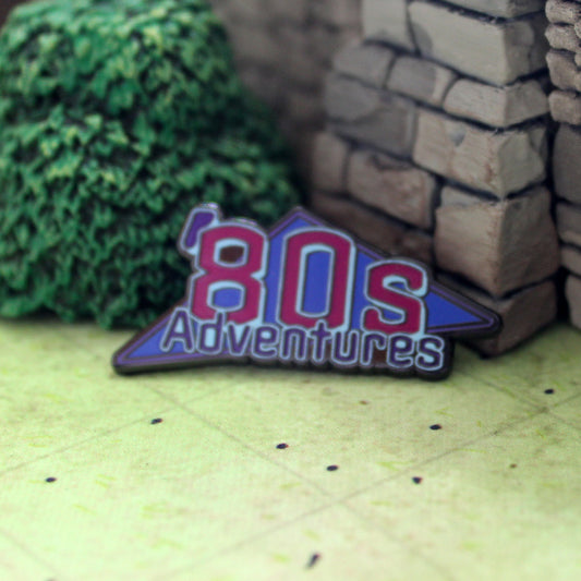 An "'80s Adventures" logo enamel pin sitting on tabletop gaming terrain
