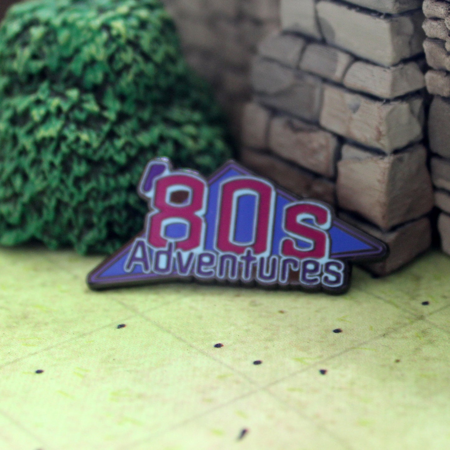 An enamel pin of an "'80s Adventures" logo sitting on tabletop gaming terrain