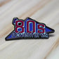 An "'80s Adventures" logo enamel pin sitting on a wooden surface.