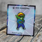 An enamel pin of a goblin wearing 1980s clothing: a red headband, sunglasses, a green jacket, blue trackpants with a red stripe, and sneakers. The goblin is holding a joystick.