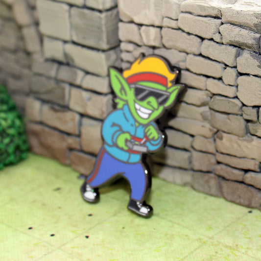 An enamel pin of a goblin wearing '80s clothing leaning against tabletop gaming terrain