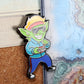 An enamel pin of a goblin wearing '80s clothing pinned to cork board