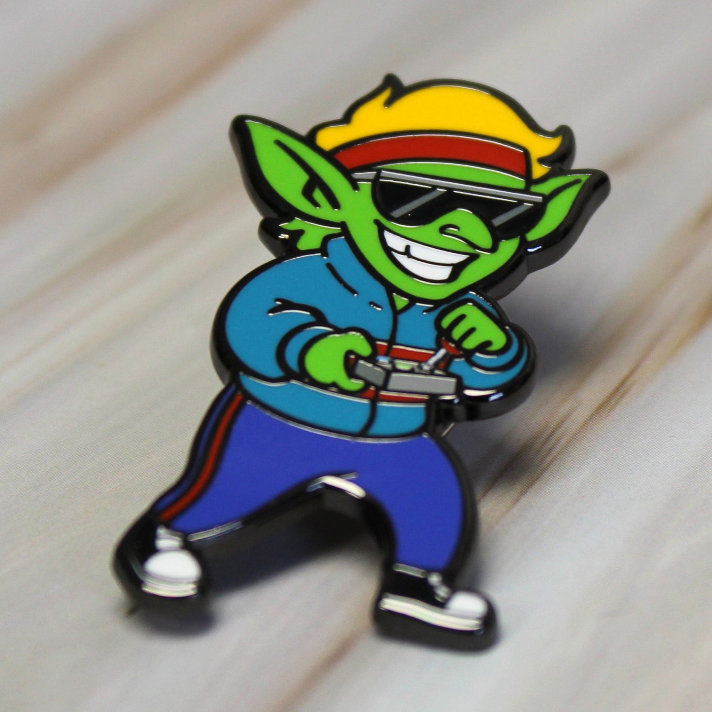 An enamel pin of a goblin wearing '80s clothing laying on a wooden surface.