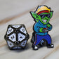 An enamel pin of a goblin wearing 1980s attire aside a twenty-sided die.