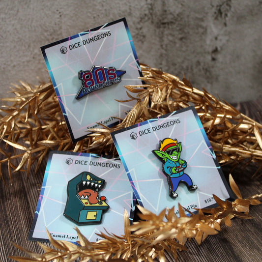 A collection of three enamel pins on neon-colored backing; one is a logo that says "'80s Adventures", one is a standup arcade machine mimic, and one is a goblin in '80s attire.