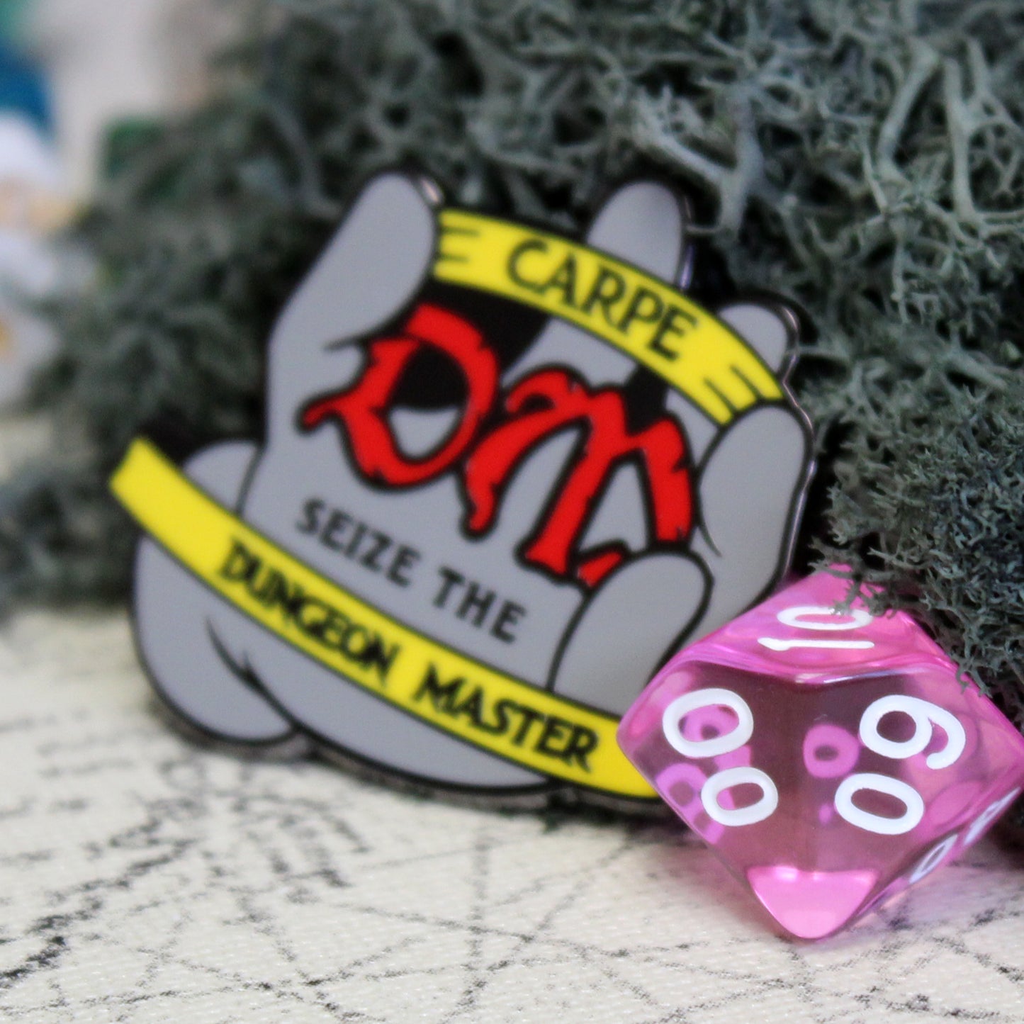 Carpe DM enamel pin placed on a tabletop beside a d% die, providing a size comparison for the accessory.