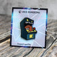 An enamel pin of a standup arcade machine mimic on a cardboard backing with a neon-colored geometric design. 