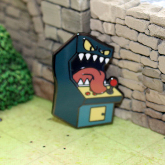 An enamel pin of a standup arcade machine mimic leaning against tabletop gaming terrain