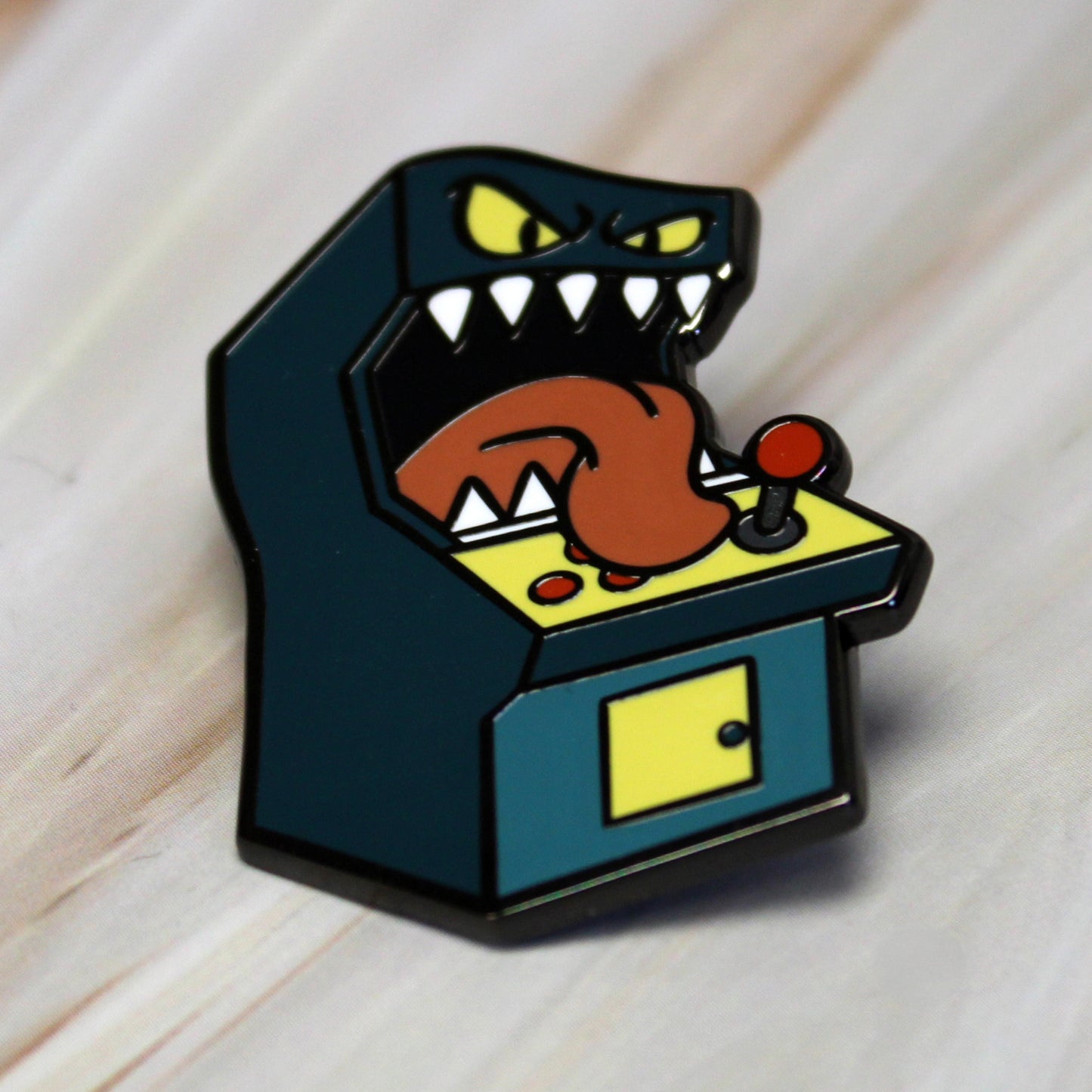 An enamel pin of a standup arcade machine mimic laying flat on a wooden surface. 